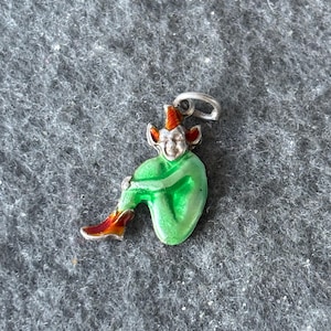 May include: A silver charm in the shape of a seated elf. The elf has a green body, red and orange hat and shoes, and a smiling face. The charm has a small loop at the top for attaching to a necklace or bracelet.