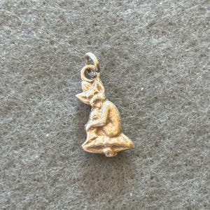 May include: A small, gold-colored charm in the shape of a seated figure, possibly a fairy or elf, with a pointed hat and flowing skirt. The charm has a loop at the top for attaching to a necklace or bracelet. The background is a textured gray fabric.