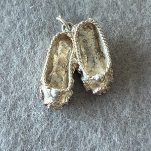 Peut inclure: Un charm en métal argenté en forme de paire de chaussons de danse. Les chaussures présentent une bordure texturée en forme de corde autour de l'ouverture et un petit détail sphérique lisse sur le bout de chaque chaussure. Un anneau est fixé au sommet.