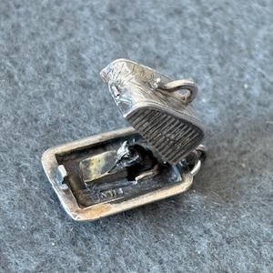 Peut inclure: Charme en argent en forme de petite boîte à charnières. La boîte est ouverte et le couvercle est relevé. Le charme a un design détaillé et est marqué "925" sur le fond.