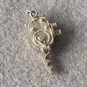 Op de afbeelding: Een zilverkleurige metalen bedel in de vorm van een vintage filmcamera. De bedel heeft een rechthoekig lichaam met cirkelvormige lensdetails, een aan de zijkant gemonteerde zwengel en een klein ringetje aan de bovenkant voor bevestiging aan een ketting.