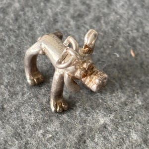 May include: A small, silver-colored dog figurine. The dog is depicted in a playful pose, with its front legs extended and its head slightly raised. The figurine has detailed features, including ears, paws, and a tail. The dog is made of a shiny metal.