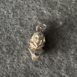 May include: A small, silver pendant depicting a sculpted bust of a person with curly hair and a ruffled collar. The pendant has a loop at the top for a chain. The bust is detailed with facial features and clothing.