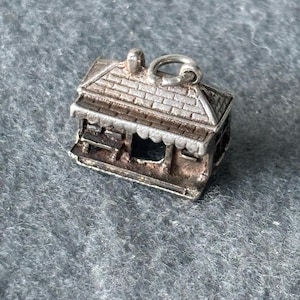 May include: A silver charm in the shape of a small house with a gabled roof and a porch. The charm has a loop at the top for attaching to a bracelet or necklace.