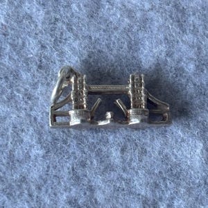 May include: A silver-toned metal charm shaped like Tower Bridge in London. The charm features two vertical towers connected by a horizontal span, with small articulated sections in the center that mimic the bridge's opening mechanism and a jump ring.