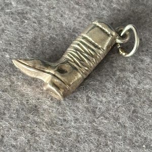 Könnte beinhalten: Ein silberfarbener Anhänger in Form eines Cowboy-Stiefels. Der Stiefel hat detaillierte Nähte und eine spitze Spitze. Oben ist eine kleine Schlaufe zum Aufhängen angebracht. Der Anhänger ist wahrscheinlich aus Metall und ein Accessoire im Western-Stil.