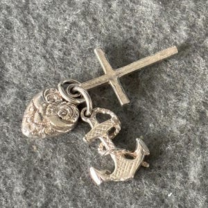 May include: A silver charm set comprising a cross, an anchor, and a heart-shaped pendant with floral detailing. The charms are linked by small rings, creating a layered effect. The background is a textured grey surface.
