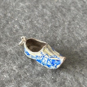 May include: A miniature silver clog charm with blue and white painted details. The small shoe has a loop for attaching to a bracelet or necklace. The clog is a traditional Dutch design.