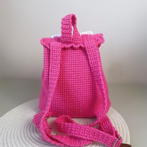 May include: A pink crocheted backpack with white trim and adjustable straps. The backpack is sitting on a white circular mat.
