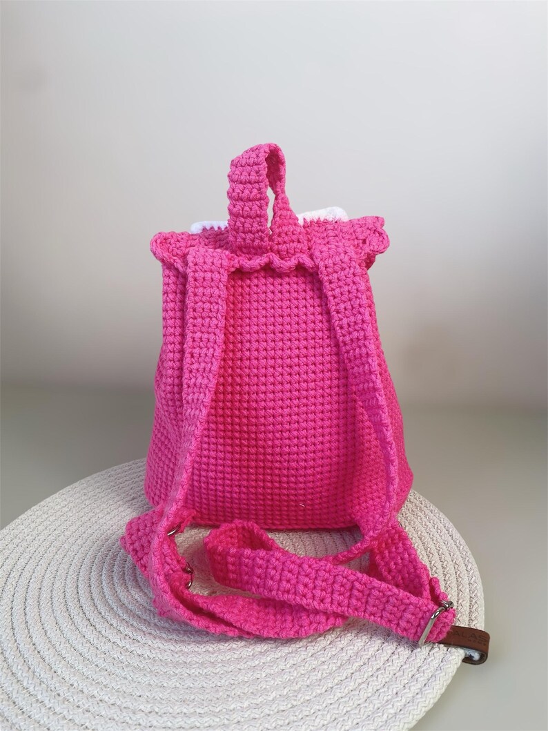 May include: A pink crocheted backpack with white trim and adjustable straps. The backpack is sitting on a white circular mat.