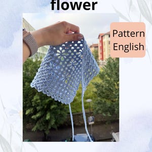Puede incluir: Se muestra un pañuelo bandana de ganchillo azul claro con un patrón de flores. El pañuelo tiene un borde festoneado y lazos largos. El texto de la imagen dice "Bandana/chal flower" y "Pattern English". También es visible el identificador de Instagram @sabinariaboutique.