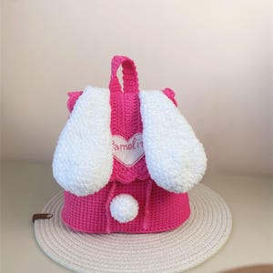 May include: Pink crocheted bunny backpack with white ears and a white pom-pom. The backpack has a heart-shaped patch with the name "Amelia" embroidered on it.