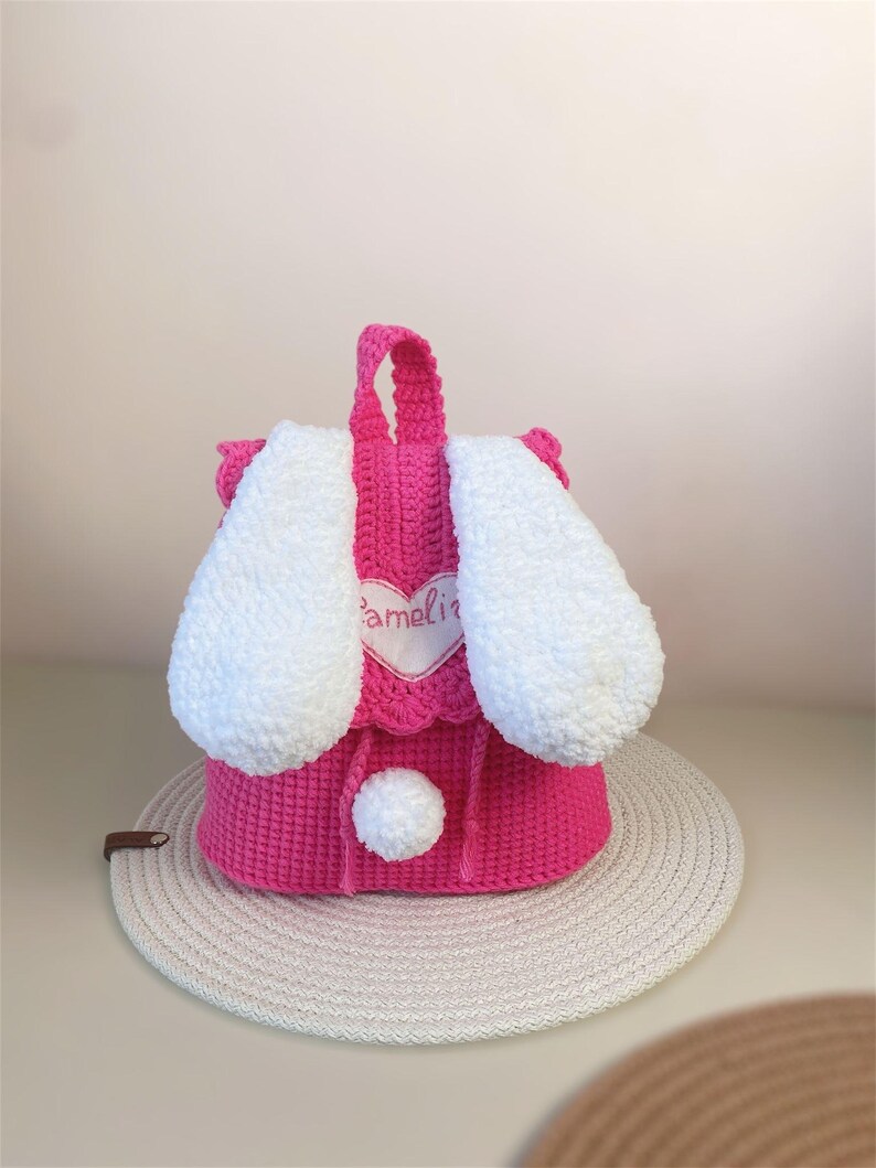 May include: Pink crocheted bunny backpack with white ears and a white pom-pom. The backpack has a heart-shaped patch with the name "Amelia" embroidered on it.