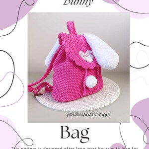 May include: Pink crocheted bunny-shaped bag with white ears and a white pom-pom. The bag has a drawstring closure and a white heart-shaped patch with the name "Camelia" embroidered on it. The bag is sitting on a woven straw mat.  @SabinariaBoutique