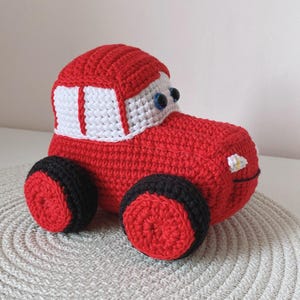 Puede incluir: Un juguete de coche rojo hecho a mano con ganchillo con ruedas negras y rojas. El coche presenta un diseño de ventana blanca y roja y ojos azules de botón. Este juguete blando es perfecto para niños.