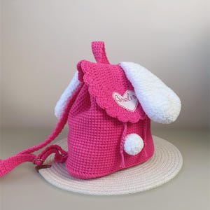 Puede incluir: Mochila de conejo de crochet rosa con orejas blancas y un pompón blanco. La mochila tiene un parche en forma de corazón con la palabra "Camelia" bordada.