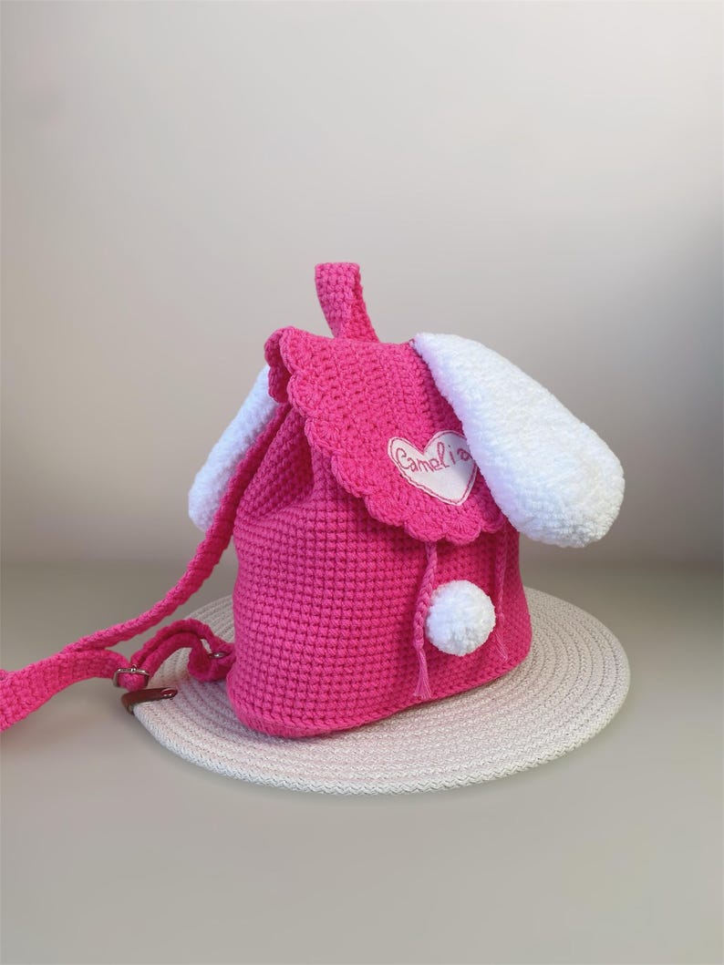 May include: Pink crocheted bunny backpack with white ears and a white pom-pom. The backpack has a heart-shaped patch with the word "Camelia" embroidered on it.