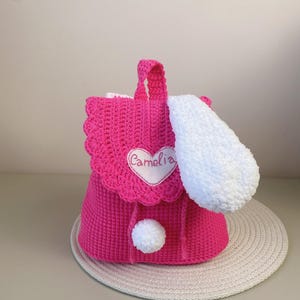 May include: A pink crocheted backpack with a white fluffy ear, a white pom-pom, and a heart-shaped patch that says "Camelia".