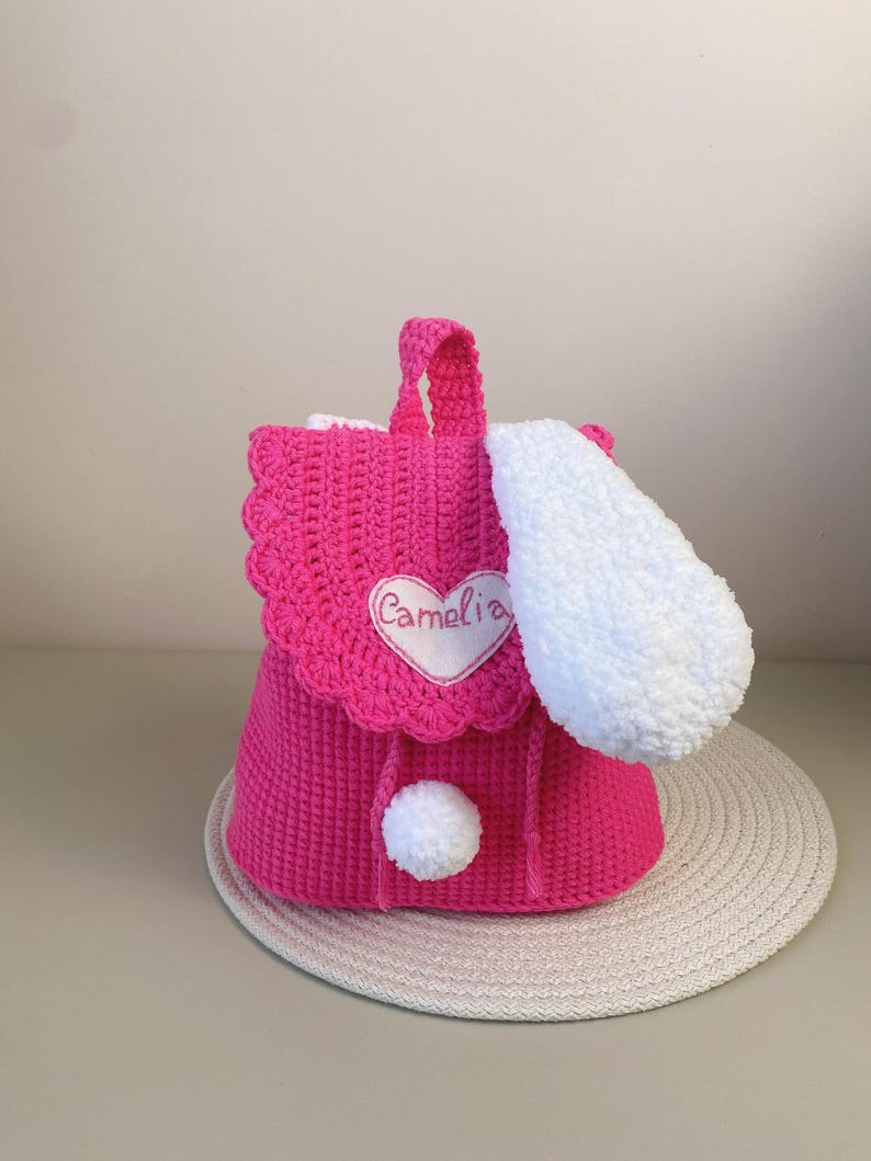 May include: A pink crocheted backpack with a white fluffy ear, a white pom-pom, and a heart-shaped patch that says "Camelia".
