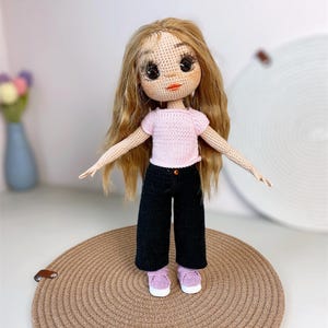 Puede incluir: Muñeca de ganchillo hecha a mano con pelo largo rubio, que lleva una camiseta rosa, pantalones negros y zapatillas lavanda. La muñeca tiene ojos grandes y expresivos y está posada sobre una estera redonda tejida.