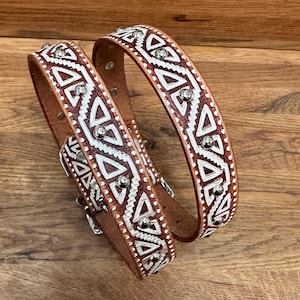 May include: A brown leather belt featuring a white geometric pattern of triangles and zig-zag lines. The belt is embellished with small, round, silver-coloured metal studs placed along the centre and edges of the strap, including a metal buckle.