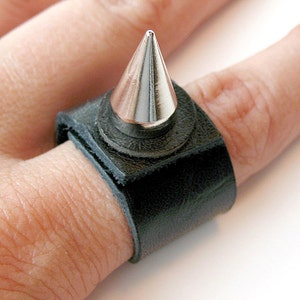 Puede incluir: Un anillo de cuero negro con una punta de plata. El anillo se lleva en un dedo.