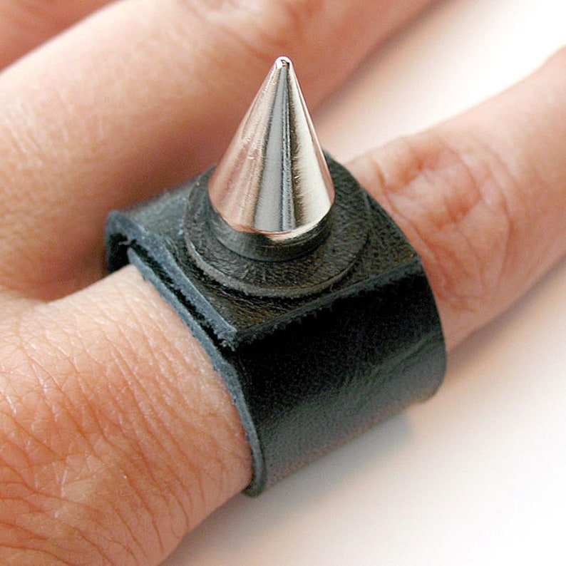 Puede incluir: Un anillo de cuero negro con una punta de plata. El anillo se lleva en un dedo.