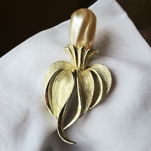 May include: A gold-toned vintage brooch with a pearl-like center and a stylized floral design. The brooch features a textured finish on the petals and leaves, creating a three-dimensional effect. The pearl-like element is elongated and has a subtle swirl pattern.