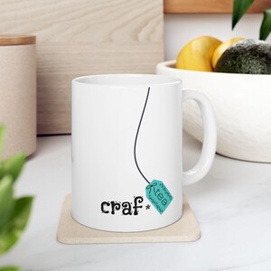 May include: White ceramic mug with a black line design and the words "craf* tea" printed on the front. The tea tag is teal with the word "tea" on it. The mug sits on a beige coaster.