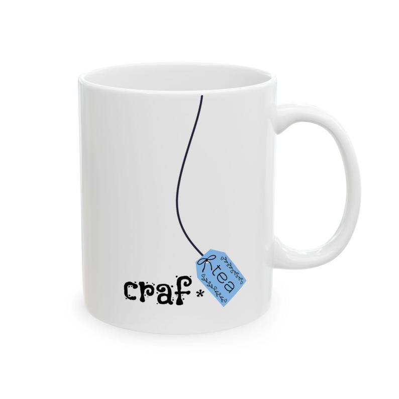 May include: White ceramic mug with a black line and a blue tea bag tag that reads "k-tea". The mug also has the word "craf*" printed on it in black, whimsical lettering. The mug has a curved handle.