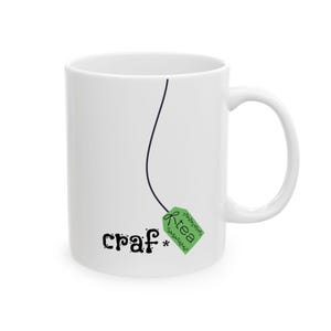 May include: White ceramic mug with a black line and a green tea bag tag design. The mug features the word "craf*" in a playful font, and the word "tea" on the tag. The mug has a curved handle.