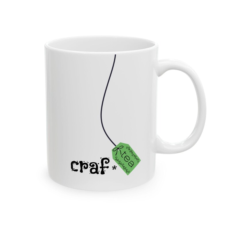 May include: White ceramic mug with a black line and a green tea bag tag design. The mug features the word "craf*" in a playful font, and the word "tea" on the tag. The mug has a curved handle.