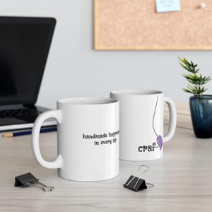 May include: Two white ceramic mugs with black text. One mug reads "handmade happiness in every sip." The other mug has the word "craft" and a purple tag design. A black laptop and binder clips are in the background.