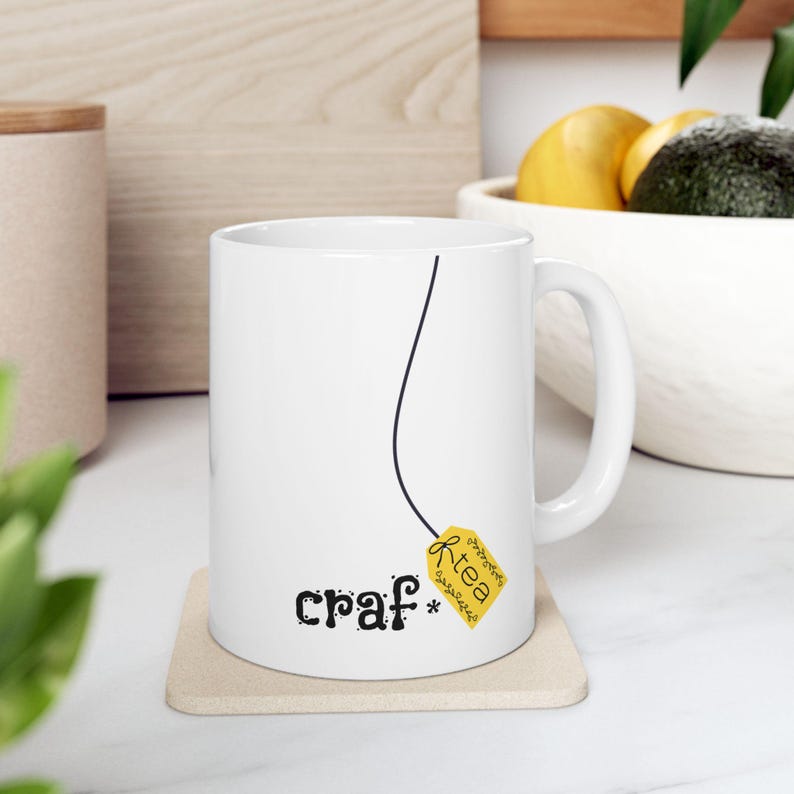 May include: White ceramic mug with a black line and a yellow tea bag tag that reads "tea". The mug also has the word "craf*" printed on it in black. The mug is sitting on a beige coaster.