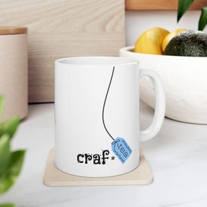 May include: White ceramic mug with a black line and a blue tea bag tag design. The mug has the word "craf*" in black script. The tea bag tag says "tea" in a decorative font. The mug is on a beige coaster.