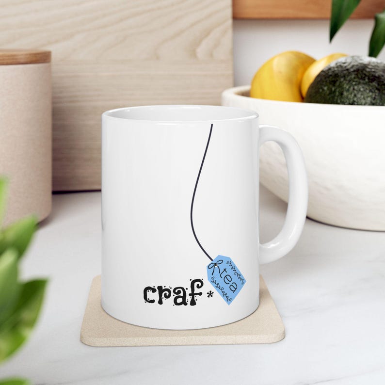 May include: White ceramic mug with a black line and a blue tea bag tag design. The mug has the word "craf*" in black script. The tea bag tag says "tea" in a decorative font. The mug is on a beige coaster.