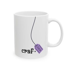 May include: White ceramic mug with a black line and a purple tea bag tag that says "rtea". The mug also has the word "craf*" in black lettering. The mug has a white handle.