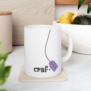 May include: White ceramic mug with a black line design and the word "craf*" in black script. A purple tea bag tag with the word "tea" is attached to the line. The mug sits on a beige coaster.