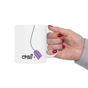 May include: White ceramic mug with the word "craf*" in a whimsical font, and a purple tea bag tag that says "tea". The mug is held by a hand with red nail polish.