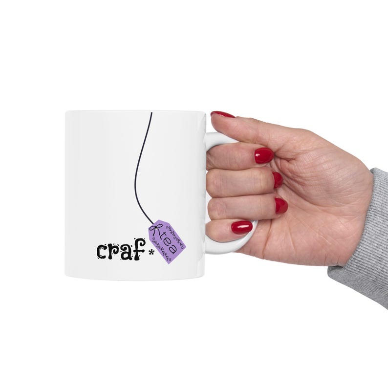 May include: White ceramic mug with the word "craf*" in a whimsical font, and a purple tea bag tag that says "tea". The mug is held by a hand with red nail polish.