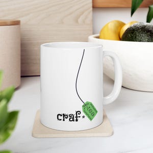May include: White ceramic mug with a black line design and the words "craf* tea" printed on the front. The tea bag tag is green with the word "tea" on it. The mug sits on a beige coaster.