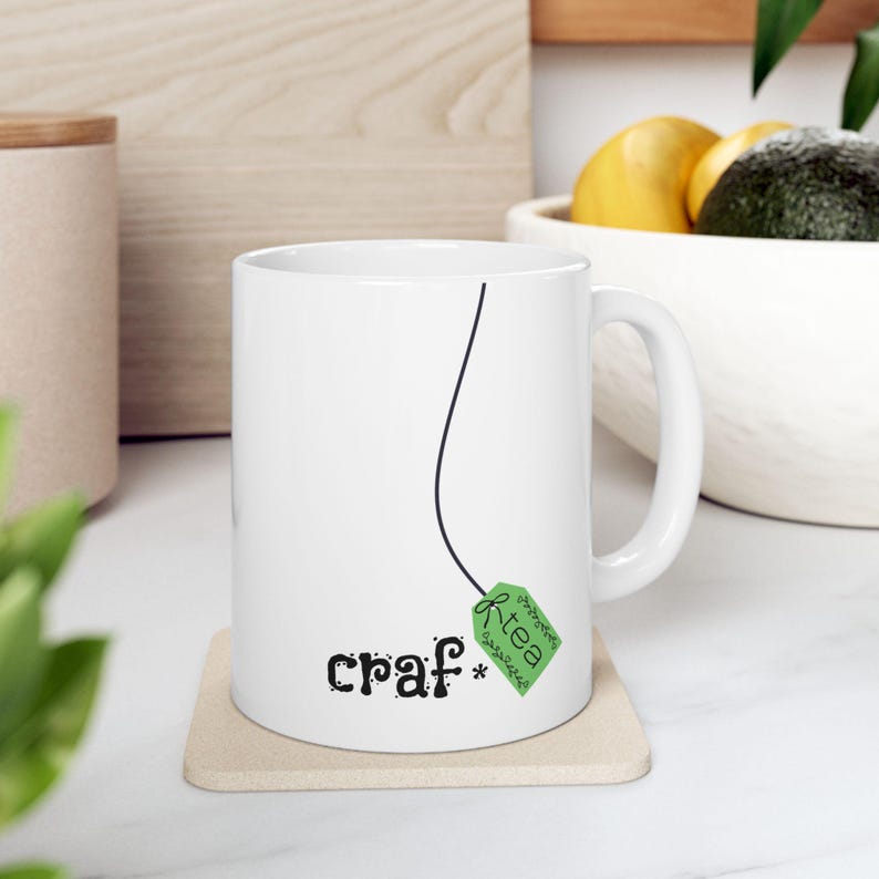 May include: White ceramic mug with a black line design and the words "craf* tea" printed on the front. The tea bag tag is green with the word "tea" on it. The mug sits on a beige coaster.