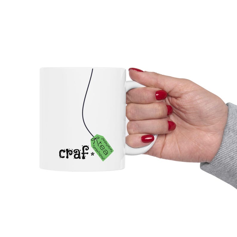 May include: White ceramic mug with the word "craf*" in black script and a green tea bag tag design. The tag reads "tea" in white script. A black line extends from the tag to the top of the mug. The mug is held by a hand with red nail polish.