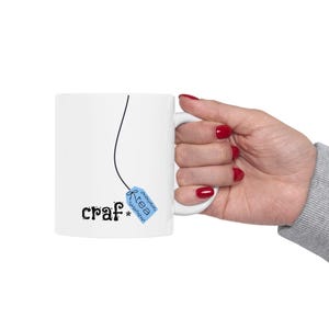 May include: White ceramic mug with the word "craf" in a whimsical font, and a blue tea bag tag that says "tea". The mug has a black string design. The mug is held by a hand with red nail polish.