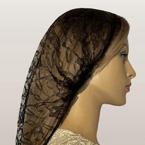 Op de afbeelding: Een zwarte kanten hoofdbedekking, gedrapeerd over het hoofd en de schouders van een mannequin. Het kant heeft een delicaat bloemmotief. De mannequin draagt een crèmekleurige kanten top. De achtergrond is neutraal.