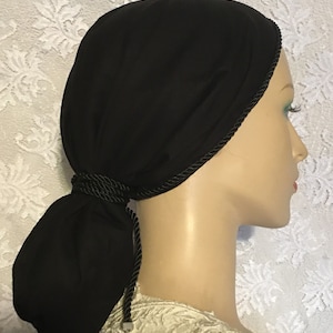 May include: Black fabric head covering with a black braided trim and a long tail that can be tied into a knot. The head covering is worn on a mannequin.