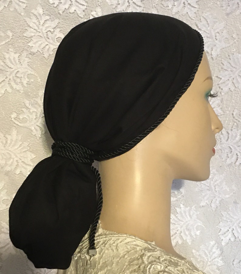 May include: Black fabric head covering with a black braided trim and a long tail that can be tied into a knot. The head covering is worn on a mannequin.