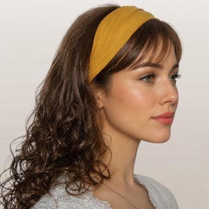 May include: A wide mustard yellow fabric headband with a gathered, ruched texture. The headband is positioned across the top of the head, resting behind the hairline and extending down towards the ears, featuring a soft, matte finish material.