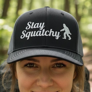 May include: Black and grey trucker hat featuring the text "Stay Squatchy" and a white Bigfoot silhouette. The hat has a mesh back and a curved peak. The wearer is smiling in a forest.