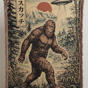 May include: Woven tapestry blanket featuring a Bigfoot illustration with a UFO, mountains, and Japanese text. The design is in shades of brown, green, and beige, with a fringed edge. The blanket is held up by a person.